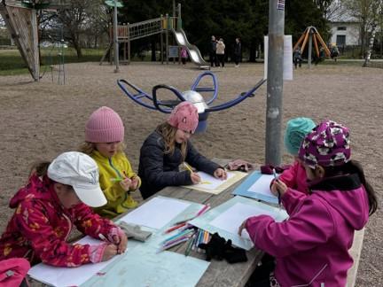Lapsia päiväkodin pihalla ulkovaatteet ja pipot päässä. Lapset istuvat ulkopöydän ääressä ja piirtävät paperille.
